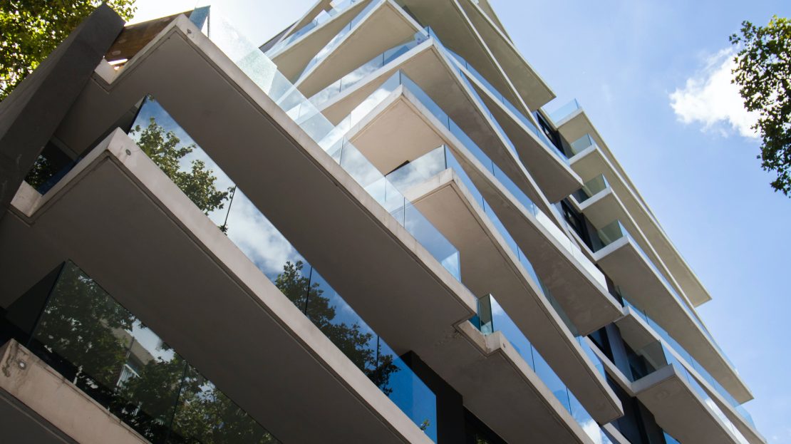 Immeuble moderne à balcons en verre vu en contre-plongée, entouré d’arbres et ciel bleu dégagé.