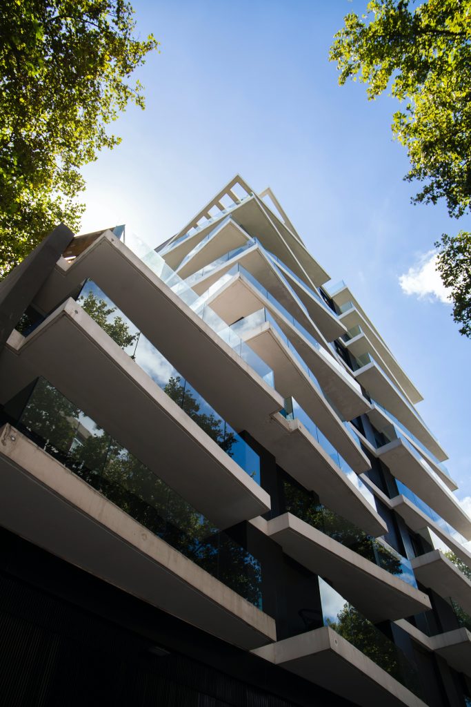 Immeuble moderne à balcons en verre vu en contre-plongée, entouré d’arbres et ciel bleu dégagé.